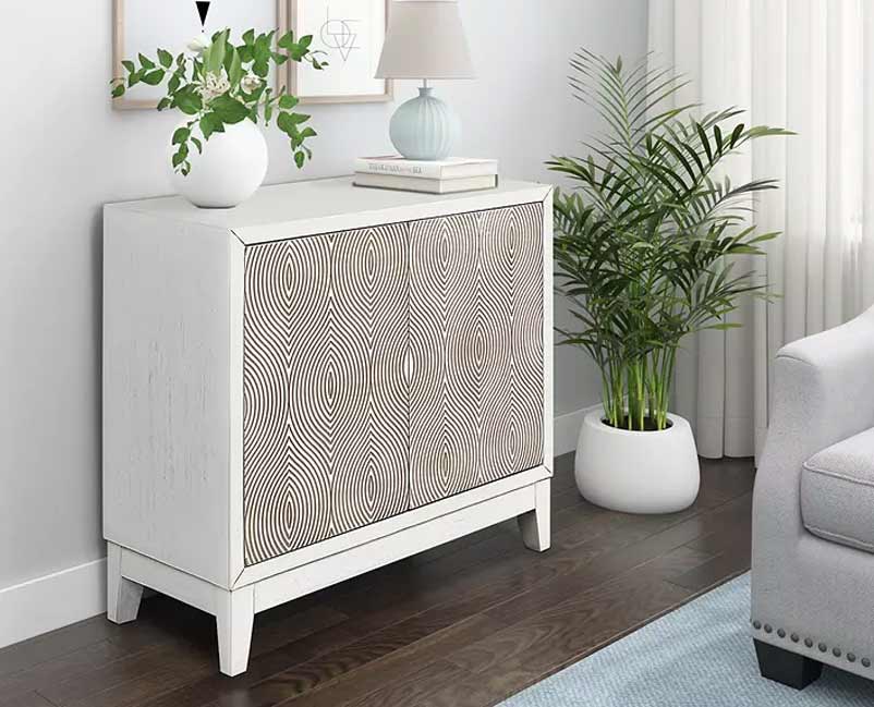Bright and airy corner of a room featuring a chic white sideboard with a unique circular patterned design, topped with a vase of fresh greenery and a soft blue lamp, adjacent to a lush potted palm plant, adding a touch of nature indoors.